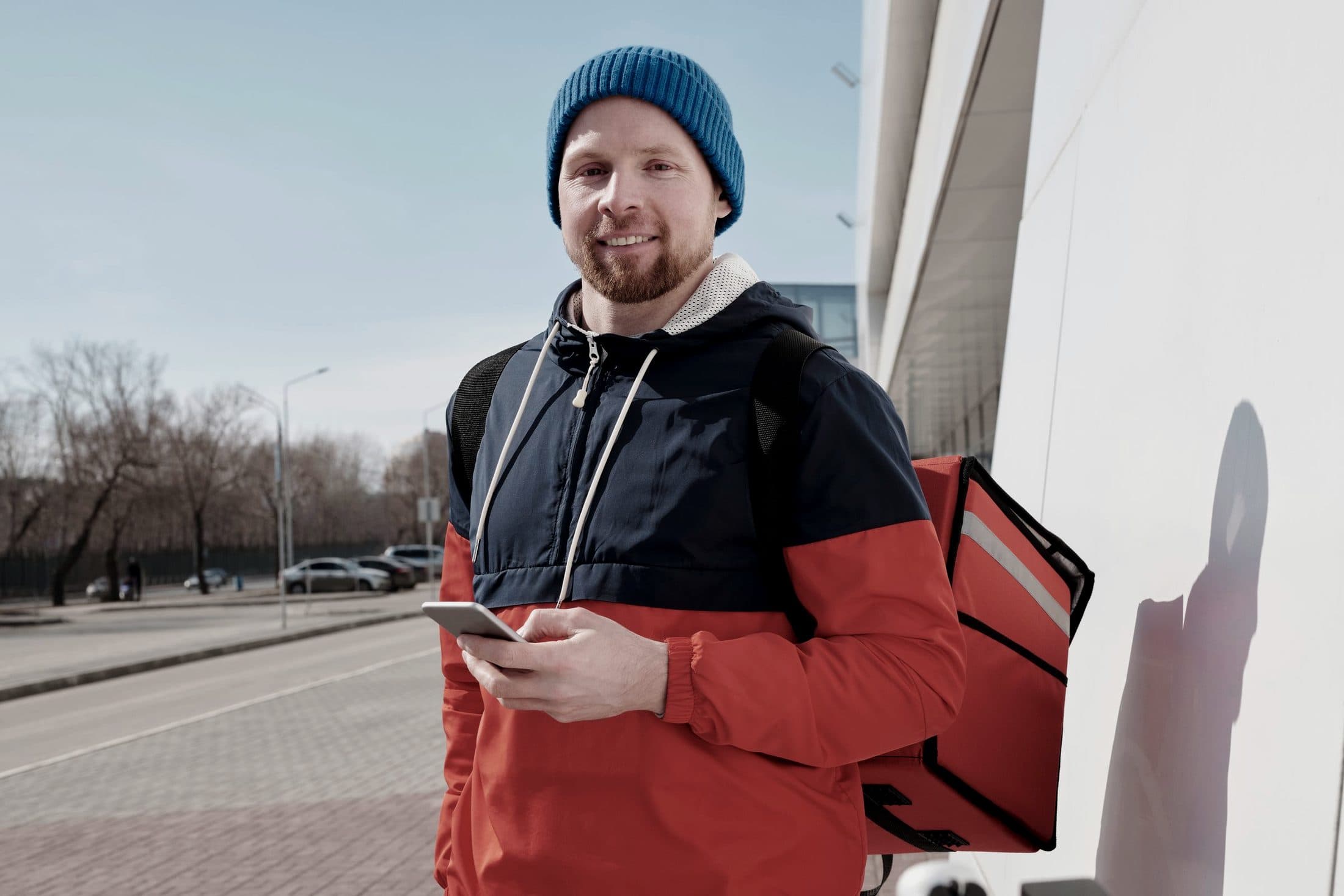 Nasmiješeni dostavljač s dostavnom torbom na leđima i mobitelom u ruci stoji na gradskoj ulici